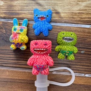 May include: A collection of colorful, whimsical monster-shaped straw toppers. The toppers are in various colors, including blue, pink, green, and yellow, with detailed facial features and patterned underwear. The toppers are attached to a white straw.