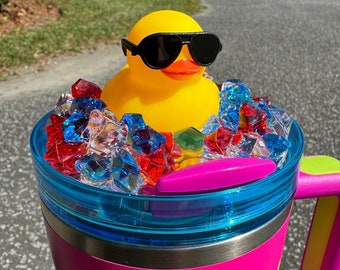 May include: A pink stainless steel travel mug with a blue lid and a rubber handle. The mug is filled with red, white, and blue candy and a yellow rubber duck wearing sunglasses.