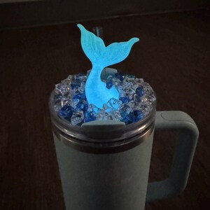 Op de afbeelding: Een turquoise beker met een handvat, bedekt met heldere en blauwe decoratieve stenen. Een gloeiende blauwe zeemeerminstaart komt uit de stenen. De beker is ontworpen om drankjes koud of warm te houden.