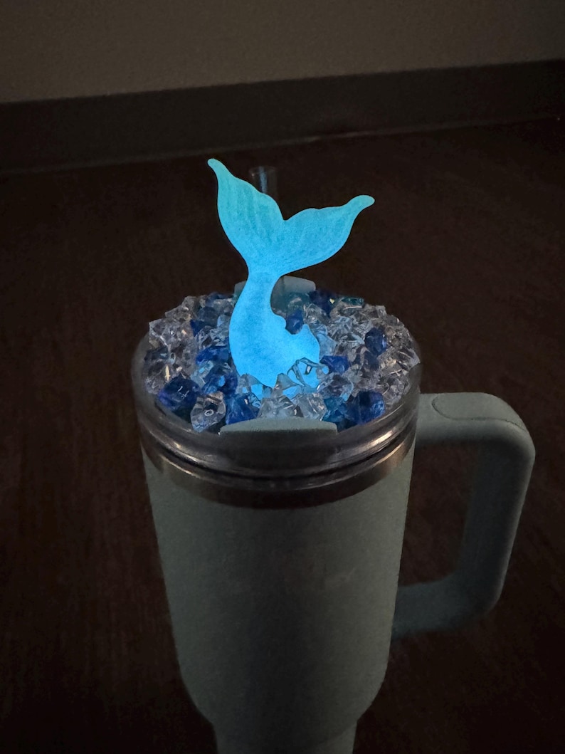 Op de afbeelding: Een turquoise beker met een handvat, bedekt met heldere en blauwe decoratieve stenen. Een gloeiende blauwe zeemeerminstaart komt uit de stenen. De beker is ontworpen om drankjes koud of warm te houden.