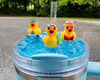 May include: A light blue tumbler with a clear lid, featuring three yellow rubber duckies with sunglasses and a hat, resting on blue ice-like material. A clear straw is inserted into the lid. The tumbler has a light blue handle.