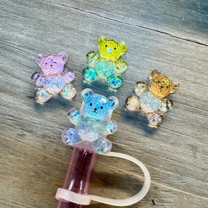 May include: Four iridescent glitter teddy bear charms in different colors: pink, yellow, blue, and brown. The charms are made of resin and have a shiny, glittery finish.