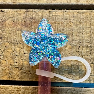 Peut inclure: Un bouchon de bouteille en forme d'étoile de mer en paillettes bleues irisées transparentes avec un élastique blanc. Le bouchon de bouteille est fixé à une bouteille en plastique transparente remplie de liquide rose.