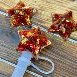 May include: Star-shaped bottle stoppers with a clear resin base, filled with red and gold leaf accents. One stopper is attached to a white straw with a loop. The stoppers are on a wooden surface.