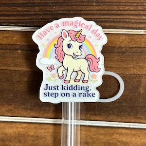 May include: A clear plastic drink stirrer with a whimsical unicorn design. The unicorn has a pink mane and tail, a yellow horn, and is set against a rainbow. The text reads "Have a magical day" and "Just kidding, step on a rake."