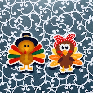 May include: Two Thanksgiving turkey figurines. One turkey is wearing a pilgrim hat and the other is wearing a red and white polka dot headband. Both turkeys have brown bodies, yellow beaks, and red wattles.
