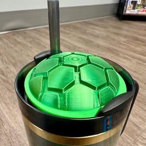 May include: A green, geometric-patterned lid sits inside a black and gold travel mug. A silver handle extends upwards from the lid. The lid has a textured, segmented design. The mug has a black exterior with a gold band near the bottom.