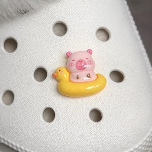 May include: A pink bear charm with closed eyes, sitting in a yellow duck floatie. The charm is attached to a white shoe with round holes. The bear has pink ears and a pink nose. The duck floatie is yellow.