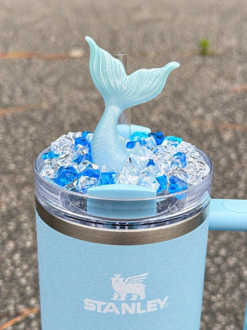 Op de afbeelding: Een lichtblauwe Stanley-beker met een zeemeerminstaart-opzetstuk. De beker heeft een doorzichtig deksel gevuld met blauwe en heldere decoratieve stenen. Het Stanley-logo is zichtbaar op de voorkant. De zeemeerminstaart is lichtblauw.