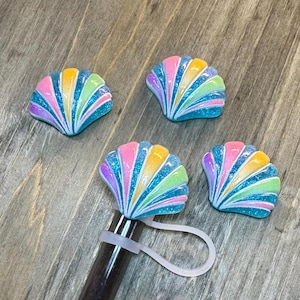 Peut inclure: Quatre embouts de stylo en forme de coquillage avec un motif arc-en-ciel. Chaque embout présente un éventail de rayures colorées en rose, jaune, vert et bleu, avec des accents de paillettes. Un embout est fixé à un stylo avec une bande de silicone blanche.