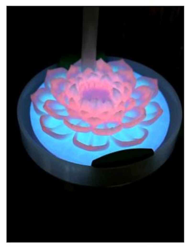 May include: A glowing lotus flower night light. The flower petals are pink and white, with a blue glow emanating from the base. The light sits in a white circular base, creating a serene and calming ambiance.