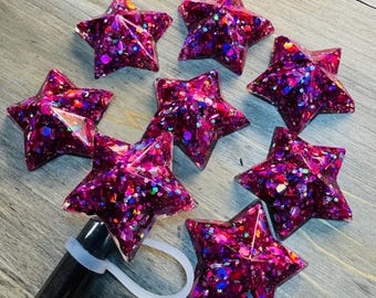 May include: Eight pink star-shaped resin ornaments with iridescent glitter embedded in the resin. The stars are arranged in a circle on a wooden surface.