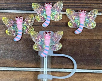 May include: Four decorative butterfly-shaped charms. Each charm features a pink body with blue stripes, glittery wings, and red eyes. One charm is attached to a clear straw with a white loop.