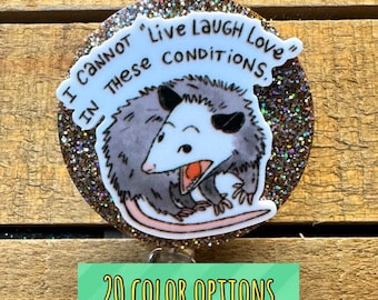 May include: A round badge with a glittery brown border features a cartoon opossum with an open mouth. The text reads "I CANNOT 'Live Laugh Love' IN THESE CONDITIONS." A green banner at the bottom says "20 COLOR OPTIONS."