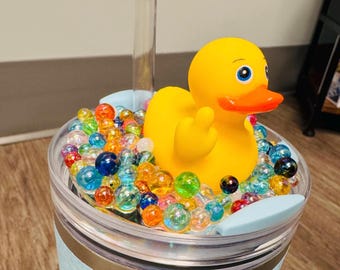 May include: A blue tumbler with a yellow rubber ducky floating in a bed of colorful beads. The tumbler has a clear lid and a straw.