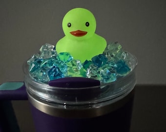 May include: A purple travel mug with a clear lid. The lid has a green rubber ducky floating in blue and green ice cubes.
