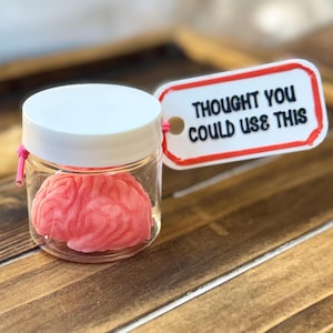 May include: A small glass jar with a white lid contains a pink brain-shaped object. A tag attached to the jar reads "THOUGHT YOU COULD USE THIS" in black text with a red border. The jar is tied with a pink string.