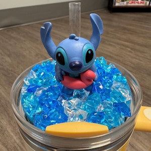 May include: A clear tumbler filled with blue and clear decorative ice. A blue Stitch figurine with a red inflatable ring is in the center. The tumbler has a yellow handle and a clear straw.