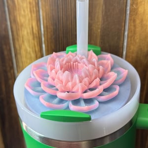 May include: A green and white insulated tumbler with a pink and blue lotus flower lid and a clear straw. The tumbler has a green handle and a silver band near the bottom. The background is a wooden wall.