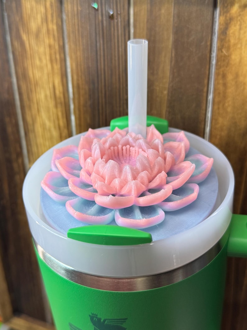 May include: A green and white insulated tumbler with a pink and blue lotus flower lid and a clear straw. The tumbler has a green handle and a silver band near the bottom. The background is a wooden wall.
