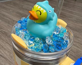May include: A yellow and white striped tumbler with a blue and green shark-shaped rubber ducky floating in blue ice crystals. The tumbler has a clear lid and a yellow straw.