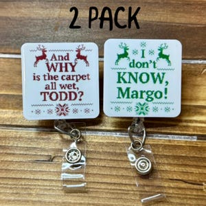May include: Two white badge reels with Christmas-themed text. One reads "And WHY is the carpet all wet, TODD?" in red. The other says "I don't KNOW, Margo!" in green. Both have reindeer and snowflake designs.