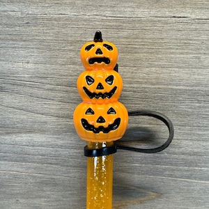 May include: A stack of three orange pumpkin-shaped Halloween decorations with black eyes and mouths. The pumpkins are attached to a black elastic band and a clear glitter-filled tube.