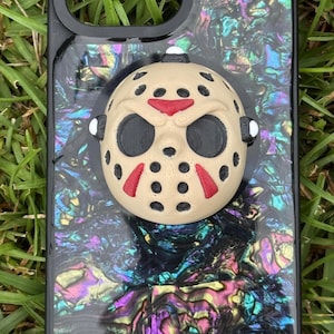 May include: A black phone case featuring an iridescent, multi-colored shell-like pattern and a three-dimensional, beige hockey mask attachment. The mask has black eye cutouts, black circular ventilation holes, and red triangular accents above the eyes.