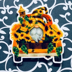 May include: A gnome wearing a yellow and black striped hat with a sunflower design sits in the back of a yellow pickup truck. The truck is filled with sunflowers and has a green and white checkered bow on the back.