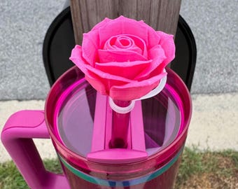May include: A pink travel mug with a matching handle and a decorative pink rose on top. The mug has a clear lid and a dark pink body with a green and blue stripe. The rose is a bright, vibrant pink.