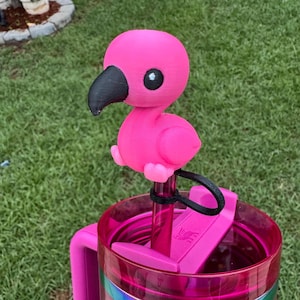 May include: A pink flamingo-shaped tumbler topper with a black beak and eye details. The topper is attached to a pink tumbler with a handle. The tumbler is sitting on green grass.