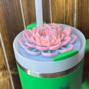 May include: A green Stanley tumbler with a white rim and a pink and blue flower-shaped lid. A clear straw extends from the lid. The tumbler has a green handle and the Stanley logo is visible on the side.