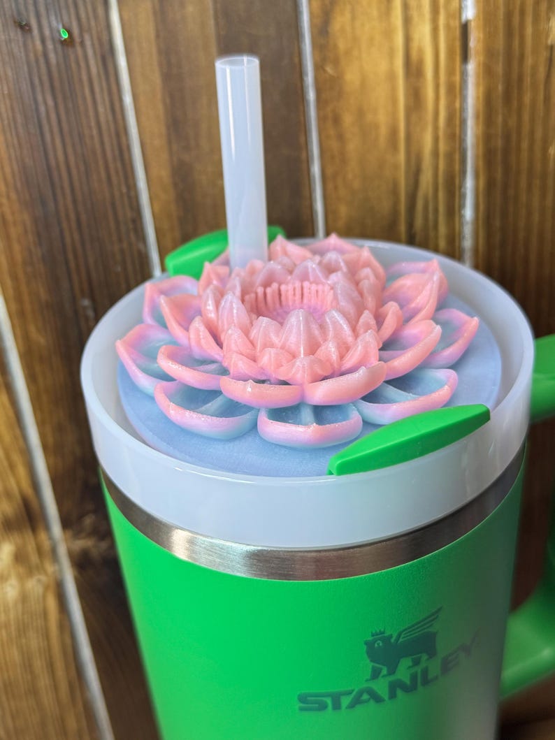 May include: A green Stanley tumbler with a white rim and a pink and blue flower-shaped lid. A clear straw extends from the lid. The tumbler has a green handle and the Stanley logo is visible on the side.