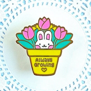 May include: A yellow enamel pin with a white bunny rabbit in a pot of pink flowers. The pot has the words "Always Growing" on it.