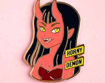 May include: A red and black enamel pin featuring a cartoon demon with horns and long black hair. The demon is wearing a red dress and has a mischievous expression. The pin has the words "Horny Demon" written on it in yellow.