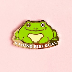 May include: A gold-colored enamel pin featuring a green frog with a yellow belly sitting on a gold background. The text "Raging Bisexual" is written in white letters on the bottom of the pin.