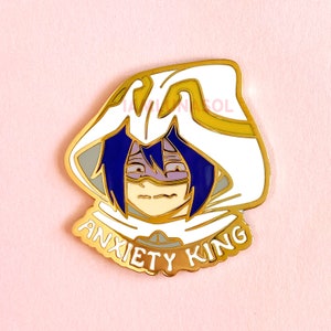 May include: Gold-colored enamel pin featuring a cartoon character with purple hair and a white hooded cloak. The character has a worried expression and is wearing a purple mask. The text "Anxiety King" is written in a gold-colored font around the bottom of the pin.