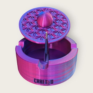 May include: A purple and blue ashtray with a flower of life design on the lid. The ashtray has a small metal poker inside and the word "CRAFT3D" is printed on the bottom.