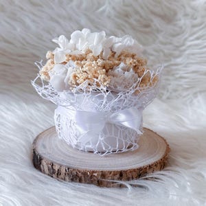 Può includere: Una piccola composizione floreale bianca in un contenitore rotondo e traforato, legata con un nastro di raso bianco. I fiori sono un mix di bianco e beige e la composizione poggia su una piccola fetta di legno rotonda.