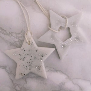 Può includere: Due scioglievoli di cera a forma di stella bianca con glitter argentato, ognuno con uno spago per appendere.
