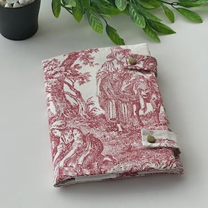 May include: A fabric-covered journal or book cover featuring a red and white toile design. The cover has a button closure and depicts pastoral scenes with figures and trees. The overall aesthetic is vintage and classic.