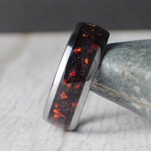 May include: A black tungsten carbide ring with a red and orange inlay. The ring is set on a gray rock.