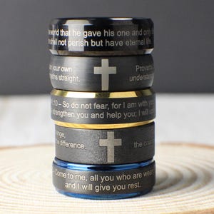 May include: A stack of five rings with engraved text and cross symbols. The rings are black, gold, and blue, with the text including Bible verses. The rings are stacked on a wooden surface.