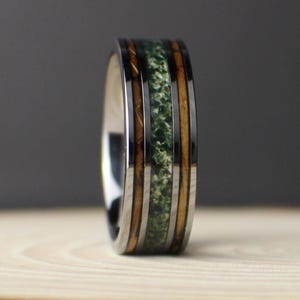 May include: A ring featuring a central band of textured green material flanked by two thin strips of brown wood, all encased within a polished dark metallic frame. The text GOODRINGS is visible in the lower left corner of the image.