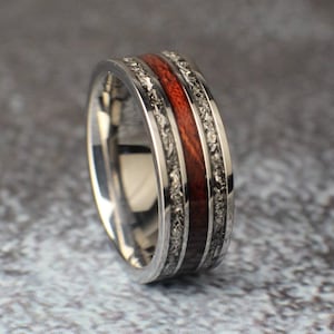 May include: A silver ring with a red wood inlay and a meteorite pattern. The ring is a wedding band.