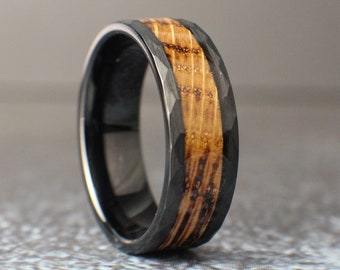 Puede incluir: Un anillo de carburo de tungsteno negro con una incrustación de madera. El anillo tiene un acabado martillado y una banda ancha.