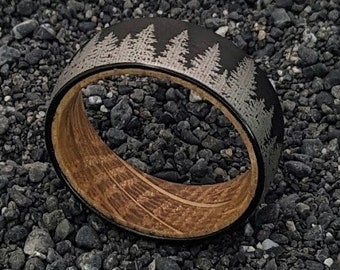 Puede incluir: Un anillo negro y plateado con una incrustación de madera y un diseño de árboles. El anillo está sobre un lecho de pequeñas piedras negras.