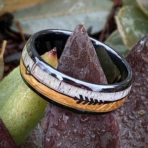 May include: A black tungsten carbide ring with a gold and white inlay featuring an arrow design. The ring is sitting on a bed of green and brown leaves.