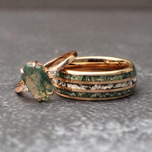 May include: Two rose gold rings. One ring features a large, green, oval-shaped stone with white accents. The other ring is a wider band with green and white inlays. The rings are set against a gray background.