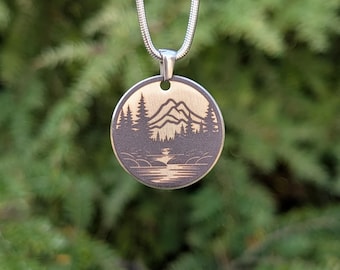 Könnte beinhalten: Eine runde Anhänger-Halskette mit einer Berg- und Waldszene. Der Anhänger ist dunkelgrau mit einem goldfarbenen Berg- und Baumdesign. Die Halskette ist silberfarben.
